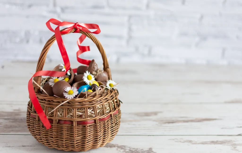 Cesta de vime cheia de ovos de pascoa de chocolate, decodada com laço vermelho na alça e pequenas margaridas decorando a cesta.